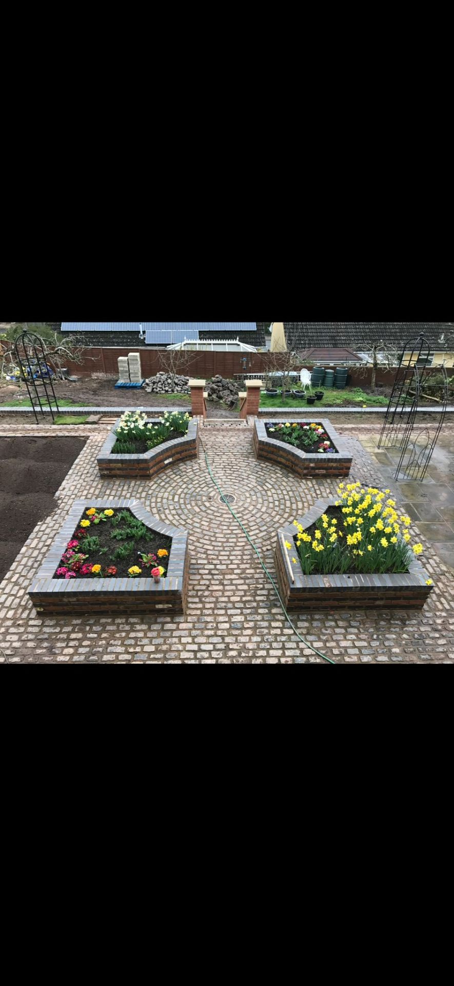 Symmetrical brick-edged formal garden with circular block paving centrepiece, raised planters of daffodils and primulas, and rose arches.