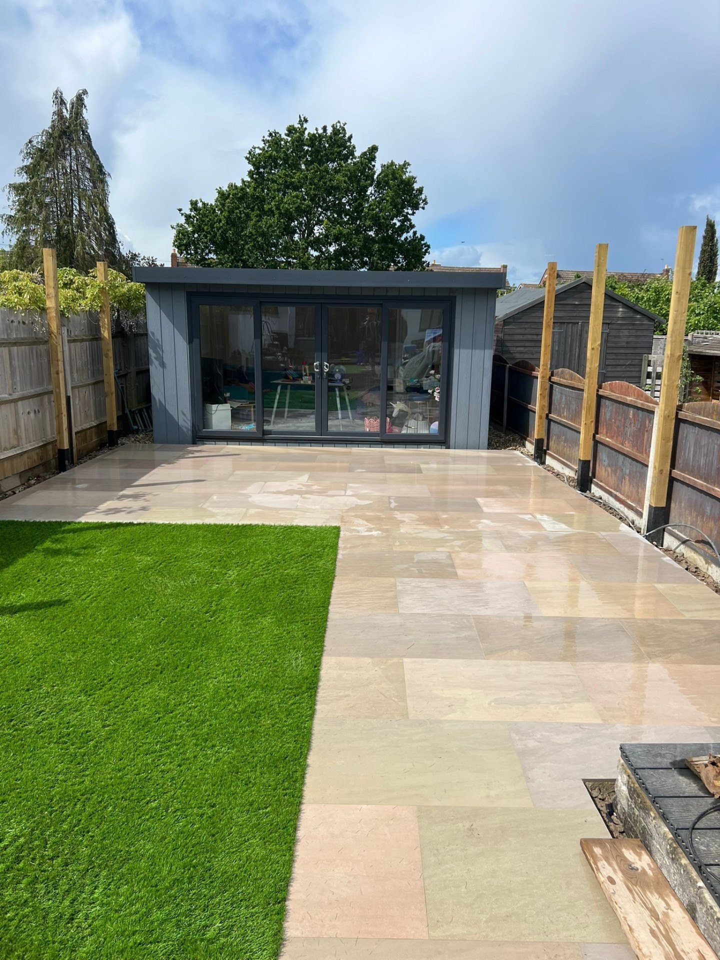 Modern garden landscape with sandstone patio flags, artificial grass, and grey summerhouse.