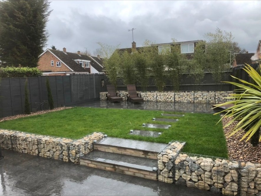 Modern garden with gabion basket retaining walls, lawn, dark paving, and bamboo screening.